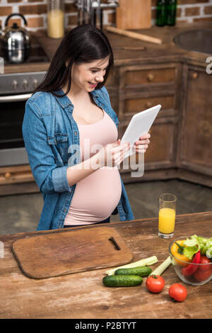 Sorridente giovane donna incinta con tavoletta digitale durante la cottura in cucina Foto Stock