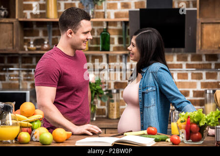 Vista laterale di felice incinta giovane coppia sorridente ogni altro mentre la cucina insieme in cucina Foto Stock