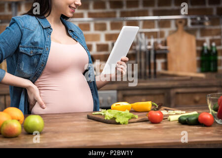 Ritagliato colpo di sorridente giovane donna incinta con tavoletta digitale durante la cottura in cucina Foto Stock