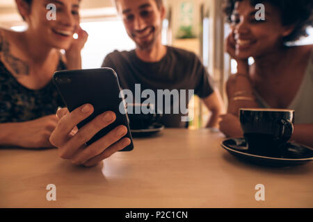 Tre giovani guardando al telefono cellulare e sorridente seduti al ristorante tavolo. Uomo che mostra qualcosa di interessante da amici di sesso femminile durante l m Foto Stock