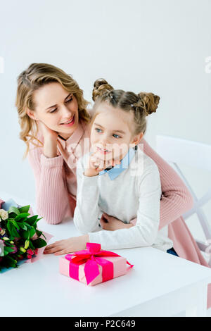 La madre abbraccia la figlia a tavola con confezione regalo e bouquet, felice festa della mamma concept Foto Stock