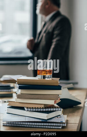 Close-up shot di bicchiere di whiskey sulla pila di libri con senior man standing sfocati sullo sfondo Foto Stock