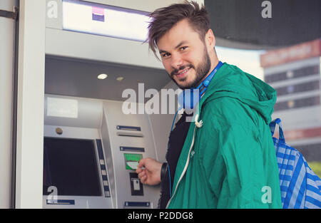 Giovani hipster guy nelle cuffie sorridente in telecamera durante l'inserimento di carta di credito nella macchina ATM ottenere denaro contante Foto Stock