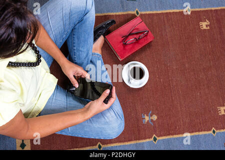 Close up della giovane donna di indossare un abbigliamento informale e collana seduti sul tappeto, tenendo un smartphone e rosso libro vintage, una tazza di caffè e blac Foto Stock