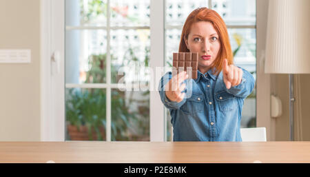 Redhead donna tenendo premuto barra di cioccolato a casa puntando con il dito per la telecamera e a voi, mano segno, positivo e fiducioso gesto dalla parte anteriore Foto Stock