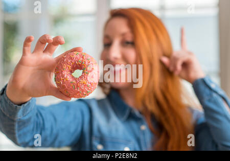 Redhead donna ciambella di contenimento a casa sorpresi con un'idea o domanda puntare il dito con la faccia felice, numero uno Foto Stock