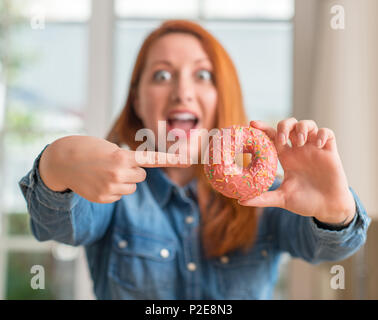 Redhead donna ciambella di contenimento a casa molto felice con puntamento della mano e del dito Foto Stock