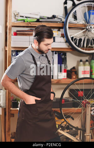 Fiducioso giovane lavoratore in grembiule guardando la ruota di bicicletta in officina Foto Stock
