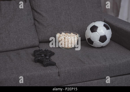 Ciotola con popcorn, sfera di calcio e gamepad sul divano Foto Stock