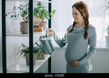 Felice donna incinta impianti di irrigazione con annaffiatoio in salotto Foto Stock