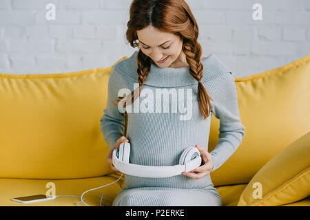 Bella donna incinta mettendo le cuffie con la musica sulla pancia in salotto Foto Stock