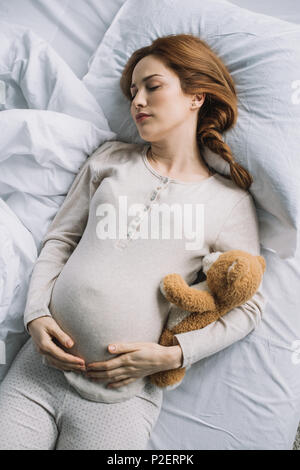 Angolo di alta vista della bella donna incinta che dorme sul letto con Teddy bear Foto Stock