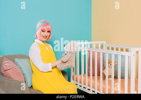 Felice in stile retrò incinta pin up donna con capelli rosa seduta con orsacchiotto vicino a culla Foto Stock