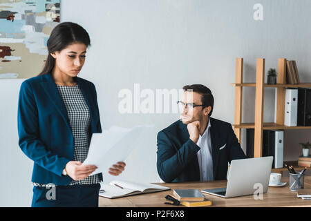 Imprenditore seduti a tavola con computer portatile mentre imprenditrice studiando contratto in ufficio moderno Foto Stock