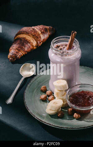 Closeup colpo di berry smoothie con cannella paglia e scaglie di cioccolato con noci, marmellata e fette di banana sulla piastra, cucchiaio e croissant Foto Stock