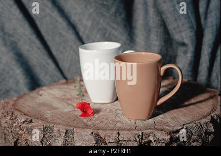 Nuovo tazze su un ceppo di legno con un fiore rosso, su un tessuto grigio Sfondo. Still Life. foto utensili Foto Stock