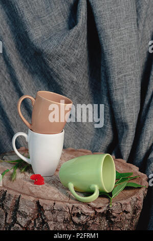 Nuovo tazze su un ceppo di legno con un fiore rosso, su uno sfondo di tessuto grigio e una scatola di legno. Ancora in vita. Foto di piatti Foto Stock