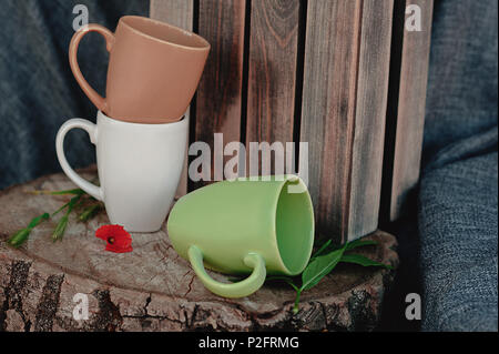 Nuovo tazze su un ceppo di legno con un fiore rosso, su uno sfondo di tessuto grigio e una scatola di legno. Ancora in vita. Foto di piatti Foto Stock