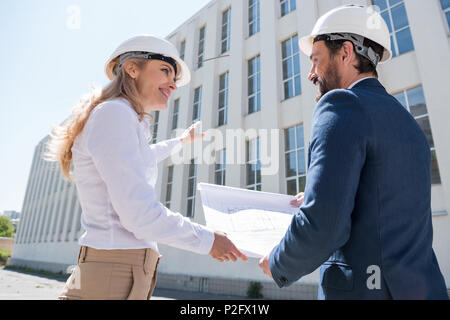 Due architetti professionisti in hardhats discutendo blueprint mentre si lavora al sito in costruzione Foto Stock