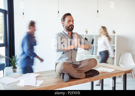 Imprenditore rilassato meditando nella posizione del loto mentre i colleghi in movimento in ufficio Foto Stock