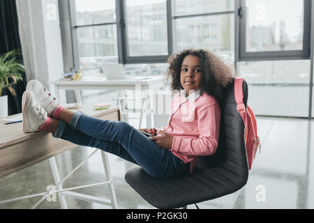 Poco americano africano ragazza seduta su una sedia con le gambe sul tavolo in ufficio moderno Foto Stock