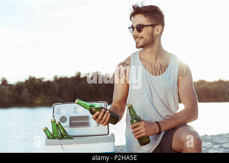 Giovane uomo informale in occhiali da sole tenendo le bottiglie di birra su Riverside Foto Stock