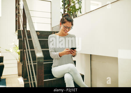 Sorridente giovane imprenditrice asiatica con gli occhiali seduto sulle scale in un ufficio la lettura dei messaggi di testo sul suo cellulare Foto Stock