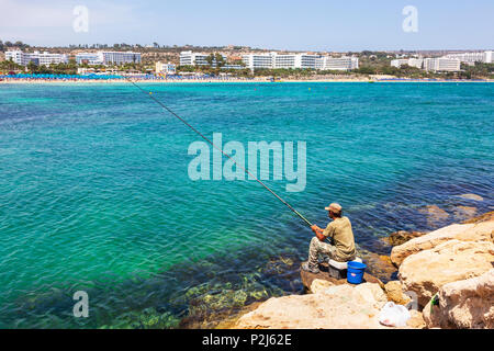 Uomo locale pesca dalla fine del faro affioramento roccioso con la località turistica di Agia Napa, Cipro a distanza Foto Stock