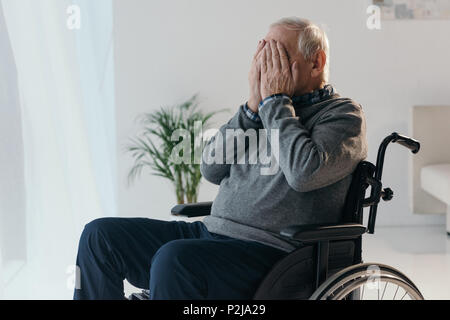 Senior triste uomo in sedia a rotelle che copre il volto con le mani Foto Stock