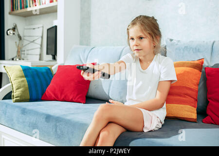 Piccola ragazza facile guardare la tv a casa. Capretto femmina seduto sul divano con il telecomando della TV e canali di commutazione Foto Stock