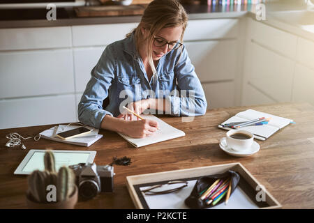 Creative giovane donna abbozzare le progettazioni con una matita su un blocco per appunti durante la seduta al suo tavolo da cucina a casa Foto Stock