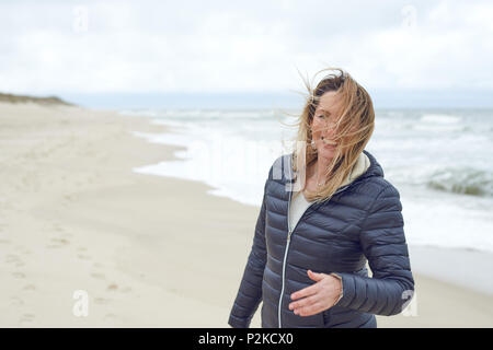 Donna sorridente su un ventoso spiaggia deserta a ridere come la brezza soffia i suoi capelli attraverso la sua faccia, con spazio di copia Foto Stock