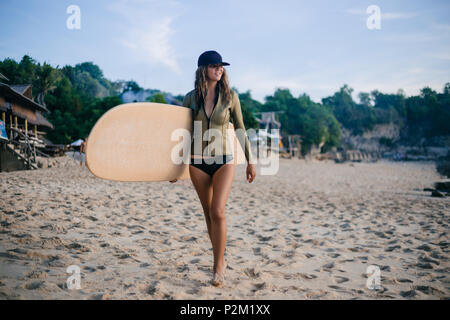 Giovane donna in wetsuit camminando dalla spiaggia con la tavola da surf in sera Foto Stock