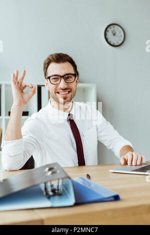 Elegante imprenditore sorridente con notebook e documenti mostra ok segno in workspace Foto Stock