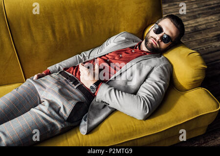 Uomo ubriaco in occhiali da sole con un bicchiere di cognac che dorme sul divano Foto Stock