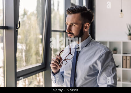 Malinconici barbuto imprenditore tenendo occhiali e guardando alla finestra in un ufficio moderno Foto Stock