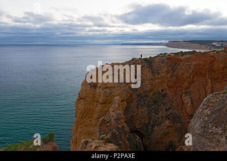 Nuvoloso Tramonto su una costa dell'Oceano Atlantico, Algarve, Portogallo. Un pescatore solitario sulla riva del litorale Scogliera al tramonto. Foto Stock