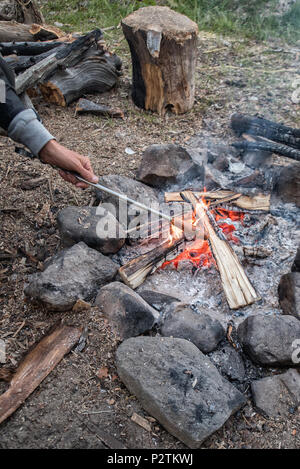 Mano d'uomo stecco di presa con una salsiccia oltre il campo di fuoco fatta sulla terra e circondato da pietre. Erba non focalizzato e legna da ardere in background. Acti Foto Stock