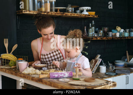 Giovane madre e figlia preparare i cookie in cucina. Essi sono in grembiuli. Bambina spruzza biscotti della pasticceria. Tempo per la famiglia. Foto Stock