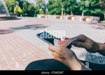 Donna di pulizia display di smart phone con antibatterico tessuto bianco, mentre sedendo nel parco pubblico. Mani femminili tenendo il dispositivo mobile e la pulizia Foto Stock