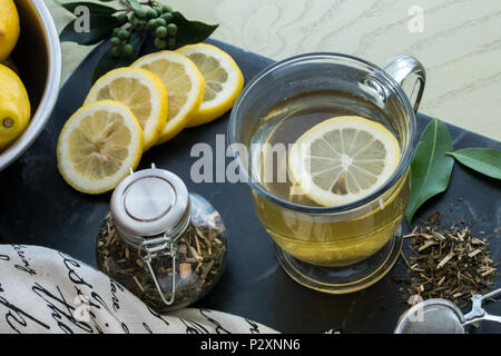 Loose Leaf Green Tea - a caldo in un vetro trasparente la tazza con una fettina di limone galleggiante sulla parte superiore della bevanda. Ciotola di limoni, tè essiccate, e disposti Foto Stock