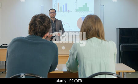 Studente maschio alzando la mano e chiedendo insegnante una questione seduta sul tavolo Foto Stock