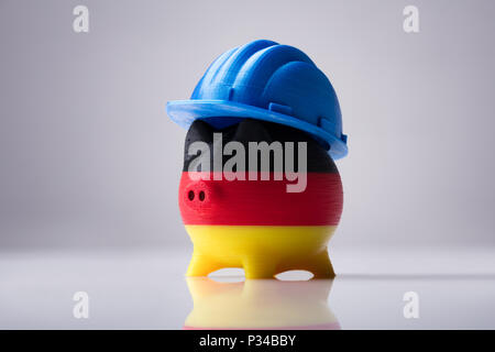 Piggybank dipinta con bandiera tedesca indossando Hardhat blu su sfondo riflettente Foto Stock