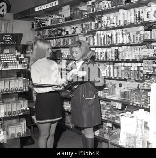 1970, due giovani donne in una farmacia, uno con i capelli lunghi e indossa una minigonna, che mostrano le altre, in un rainmac, la gamma di cosmetici e prodotti per capelli disponibile, Inghilterra, Regno Unito. Foto Stock