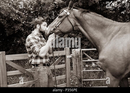 Guardando bene, hunky cowboy sorge accanto al ranch petting gate e di amare il suo cavallo Foto Stock