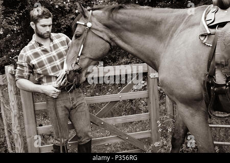 Guardando bene, hunky cowboy sorge accanto al ranch petting gate e di amare il suo cavallo Foto Stock