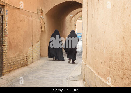Yazd, Iran - Marzo 23, 2018: due donne musulmane, vestito di nero chador a piedi sulla strada stretta del vecchio adobe Yazd città Foto Stock