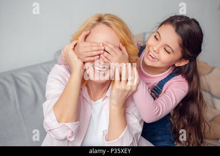 Bambina e donna senior a casa celebrazione insieme seduti sul divano ragazza che copre gli occhi della nonnina giocoso rendendo sorpresa sorridente allegro vicino- Foto Stock