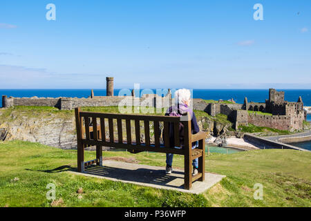 Donna anziana in pensione OAP signora rilassarsi da sola in vacanza seduta su una panchina su una collina che domina le rovine del castello di Peel. Peel, Isola di Man, Isole britanniche Foto Stock