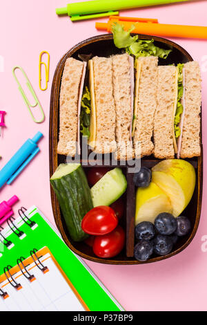 Lay piatto di legno con scatola di pranzo con panini, frutta e verdura su sfondo rosa e forniture scolastiche. Bambini sani mangiare concetto. Top vie Foto Stock